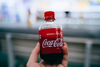 A hand is holding a small plastic bottle of Coca-Cola with a red label prominently displaying the brand name. The background is blurred, focusing attention on the bottle.
