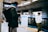 A person with a backpack is standing on a train platform, looking at their phone. The setting appears to be indoors, with fluorescent lighting and a modern, urban design. Another person is visible in the background, wearing a white sweater and green skirt.