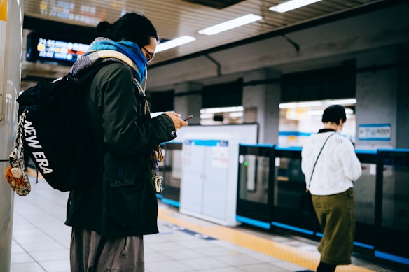 A person with a backpack is standing on a train platform, looking at their phone. The setting appears to be indoors, with fluorescent lighting and a modern, urban design. Another person is visible in the background, wearing a white sweater and green skirt.