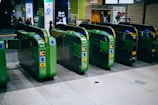 Several automatic ticket gates are lined up at a train station entrance, predominantly green in color with visible IC card slots and directional arrows. In the background, there are posters on the walls and a few people walking past.