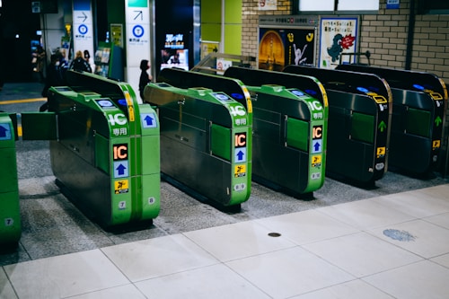 Several automatic ticket gates are lined up at a train station entrance, predominantly green in color with visible IC card slots and directional arrows. In the background, there are posters on the walls and a few people walking past.