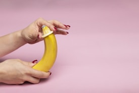 A hand with red-painted nails is holding a banana and demonstrating the application of a condom. The background is a soft pink color.