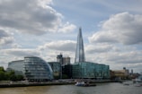 A cityscape featuring modern architecture with a prominent glass skyscraper, known as The Shard, surrounded by other contemporary and older buildings along a riverbank. The sky above is partly cloudy, adding contrast to the urban scene.