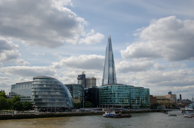 A cityscape featuring modern architecture with a prominent glass skyscraper, known as The Shard, surrounded by other contemporary and older buildings along a riverbank. The sky above is partly cloudy, adding contrast to the urban scene.