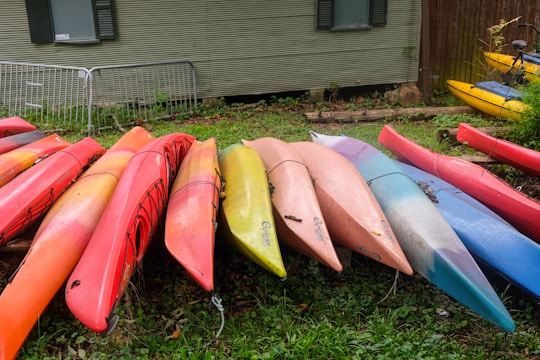 A group of colorful kayaks are arranged in a row on a grassy surface. The kayaks are primarily shades of red, orange, pink, yellow, and blue. They are stacked next to a building with corrugated metal siding, which has a window and a fence in the background. The area appears to be outdoors, adjacent to a small patch of vegetation.