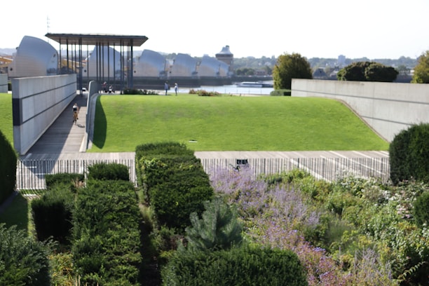 A landscaped garden with neatly trimmed hedges and various flowering plants in the foreground. In the background, an expansive green lawn stretches up to a modern architectural structure with angular, metallic elements. A pathway bisects the lawn, with a person walking along it. Trees and a river are visible further back, under a clear sky.