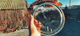 A hand is holding a round, translucent disc of ice in an outdoor setting. In the background, there is a structure with a corrugated iron roof, some dried vines and a paved area. The ice disc exhibits unique patterns formed by freezing.