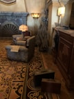 A vintage-style hotel lobby with ornate furnishings and decor. There is a patterned rug on the floor, two upholstered armchairs with a small table between them, and a newspaper resting on one armchair. The room includes a fireplace with intricate details and several floor lamps. A pair of old-fashioned suitcases are placed on the floor, and wardrobe items are visible on a nearby rack.