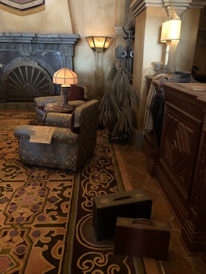 A vintage-style hotel lobby with ornate furnishings and decor. There is a patterned rug on the floor, two upholstered armchairs with a small table between them, and a newspaper resting on one armchair. The room includes a fireplace with intricate details and several floor lamps. A pair of old-fashioned suitcases are placed on the floor, and wardrobe items are visible on a nearby rack.