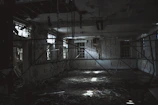 A shadowy corridor inside a vacant building with scattered debris and light filtering through broken windows.