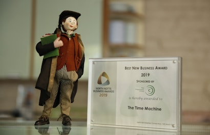 A small figurine dressed in a colorful outfit stands on a surface next to a plaque awarding the 'Best New Business Award 2019' to 'The Time Machine'. The figurine wears a long coat, striped pants, and a hat, holding a book or a piece of cloth. The award is displayed in a transparent frame with a logo and text indicating the sponsor.