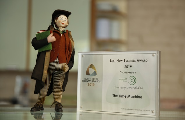 A small figurine dressed in a colorful outfit stands on a surface next to a plaque awarding the 'Best New Business Award 2019' to 'The Time Machine'. The figurine wears a long coat, striped pants, and a hat, holding a book or a piece of cloth. The award is displayed in a transparent frame with a logo and text indicating the sponsor.