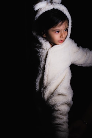 A young child wearing a white, fuzzy bunny costume stands against a dark background. The costume's hood features bunny ears, and the child appears to be looking off to the side with a curious expression.