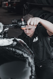 A person wearing a black shirt is actively working on a motorcycle, focusing on a task with determination. The background contains tires stacked in a garage environment.