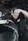 A person wearing a black shirt is actively working on a motorcycle, focusing on a task with determination. The background contains tires stacked in a garage environment.