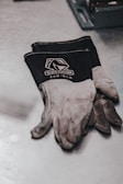 A pair of black and gray work gloves with the brand name 'Black Stallion' and the model '25D-BLK' printed on them are resting on a metal surface. Next to the gloves, a metal tool tray is partially visible.