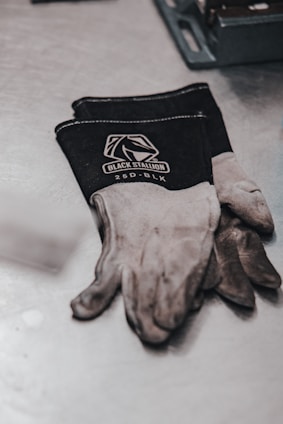 A pair of black and gray work gloves with the brand name 'Black Stallion' and the model '25D-BLK' printed on them are resting on a metal surface. Next to the gloves, a metal tool tray is partially visible.
