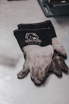 A pair of black and gray work gloves with the brand name 'Black Stallion' and the model '25D-BLK' printed on them are resting on a metal surface. Next to the gloves, a metal tool tray is partially visible.