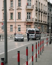 A street scene featuring a police van and a car stopped by officers who have set up a barrier with red and white tape. The buildings in the background are residential, with multiple windows and a small balcony. The atmosphere appears calm yet organized, as the officers direct traffic.