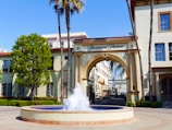 An ornate entrance archway labeled 'Paramount Pictures' stands prominently. The arch is flanked by tall palm trees and a lush green tree on one side. In front of the arch is a fountain with a clear blue water display. Surrounding the fountain is a neatly maintained garden with several windows in the building visible, suggesting a sunny day.
