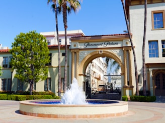 An ornate entrance archway labeled 'Paramount Pictures' stands prominently. The arch is flanked by tall palm trees and a lush green tree on one side. In front of the arch is a fountain with a clear blue water display. Surrounding the fountain is a neatly maintained garden with several windows in the building visible, suggesting a sunny day.
