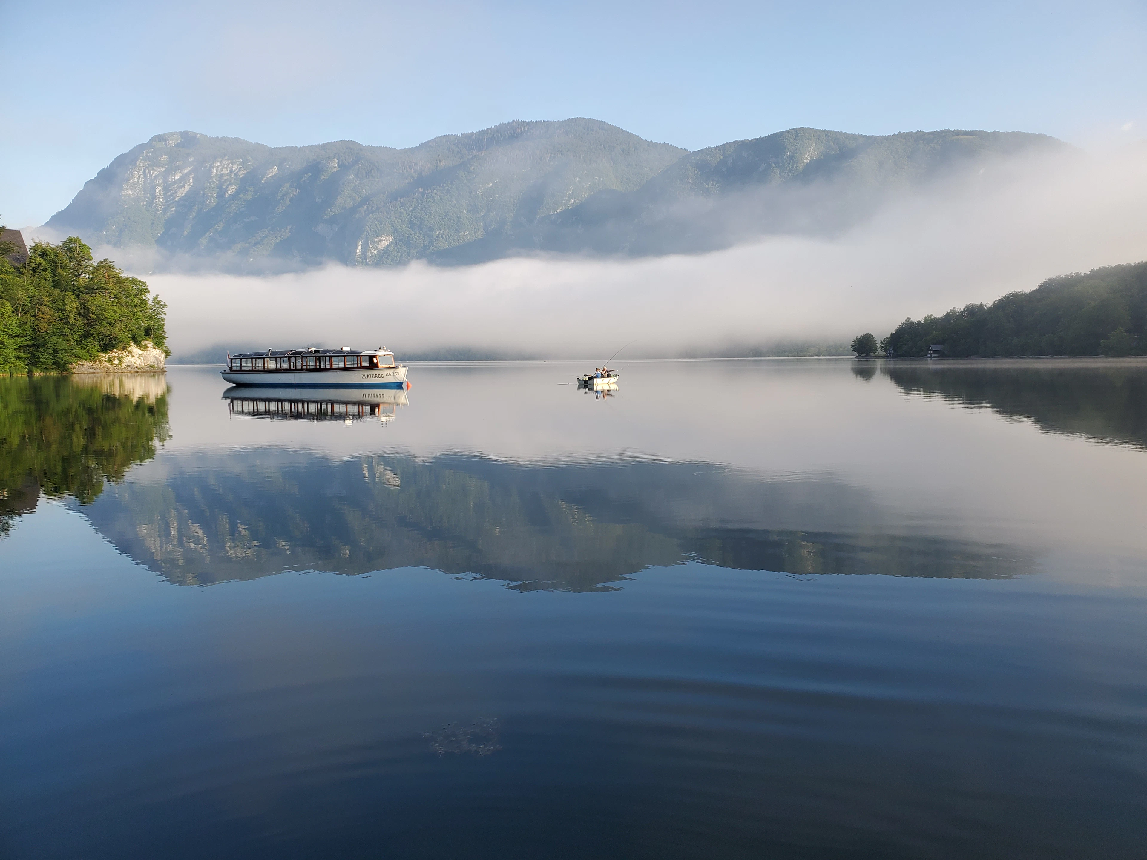 Ryto rūkas ir veidrodinis atspindys Bohinjo ežere, Slovėnijoje: prie kranto stovi pramoginis laivas, toliau – nedidelė valtis