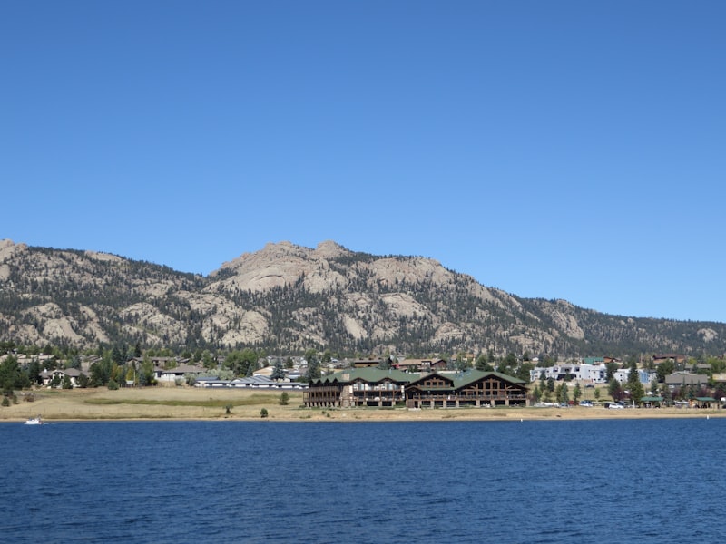 Deep blue lake with a small sailboat, a timber lodge on the shore and the rounded granite domes of Lumpy Ridge rising behind a hillside of houses under a clear sky