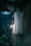 A ghostly figure hangs from the ceiling in a dimly lit space with brick walls. The figure appears to be a doll with a white sheet covering it and is positioned next to a hanging light fixture. Below it, a mannequin leg with a painted red toenail dangles. There is a potted plant partially visible at the bottom left corner.