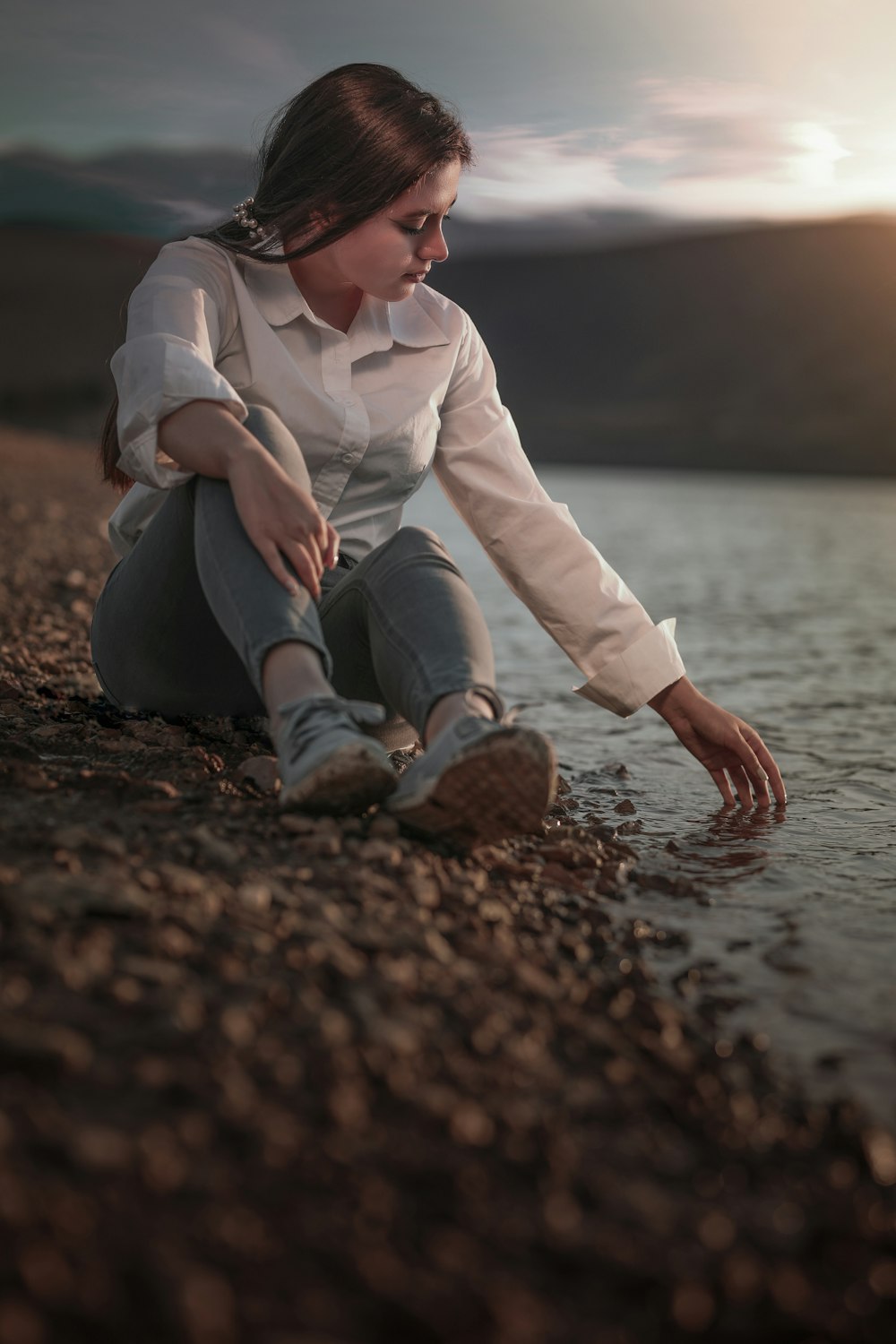 woman in white dress shirt and blue denim jeans sitting on brown rock near body of on on on on