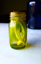 A glass jar filled with pickled vegetables, primarily green in color, is placed on a table. The jar contains long, sliced pieces submerged in liquid, with a sealed lid on top. In the background, a blurred dark blue or black object is partially visible.