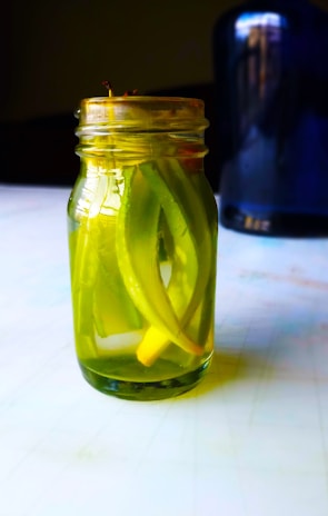 A glass jar filled with pickled vegetables, primarily green in color, is placed on a table. The jar contains long, sliced pieces submerged in liquid, with a sealed lid on top. In the background, a blurred dark blue or black object is partially visible.