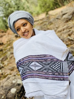 A person wearing a traditional white shawl with intricate black and purple patterns stands outside. They are also wearing a gray head wrap and are smiling. The background is a natural setting with rocks and greenery, suggesting an outdoor location.