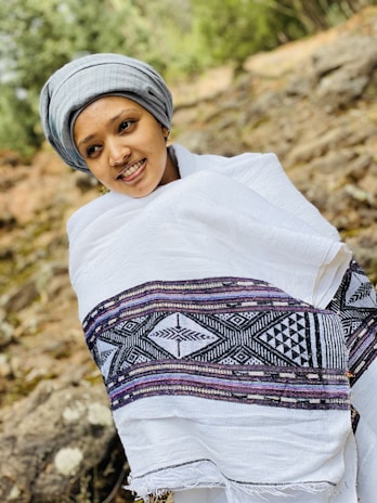 A person wearing a traditional white shawl with intricate black and purple patterns stands outside. They are also wearing a gray head wrap and are smiling. The background is a natural setting with rocks and greenery, suggesting an outdoor location.