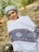 A person wearing a traditional white shawl with intricate black and purple patterns stands outside. They are also wearing a gray head wrap and are smiling. The background is a natural setting with rocks and greenery, suggesting an outdoor location.