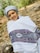 A person wearing a traditional white shawl with intricate black and purple patterns stands outside. They are also wearing a gray head wrap and are smiling. The background is a natural setting with rocks and greenery, suggesting an outdoor location.