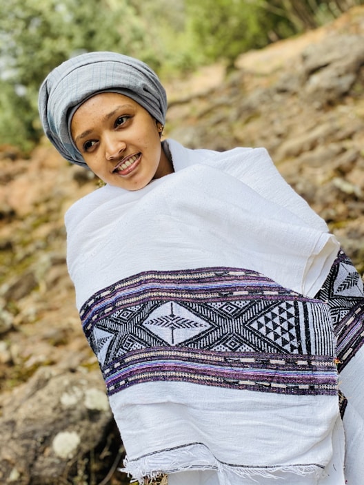A person wearing a traditional white shawl with intricate black and purple patterns stands outside. They are also wearing a gray head wrap and are smiling. The background is a natural setting with rocks and greenery, suggesting an outdoor location.
