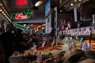 A bustling indoor market with a seafood stall displaying a variety of fresh seafood on ice. Neon signs hang above, including one for a restroom and another indicating fresh products. People are walking around and examining the goods.