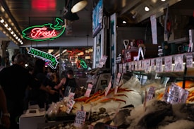 A bustling indoor market with a seafood stall displaying a variety of fresh seafood on ice. Neon signs hang above, including one for a restroom and another indicating fresh products. People are walking around and examining the goods.