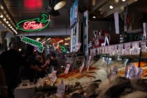 A bustling indoor market with a seafood stall displaying a variety of fresh seafood on ice. Neon signs hang above, including one for a restroom and another indicating fresh products. People are walking around and examining the goods.