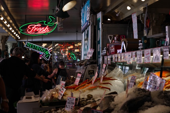 A bustling indoor market with a seafood stall displaying a variety of fresh seafood on ice. Neon signs hang above, including one for a restroom and another indicating fresh products. People are walking around and examining the goods.