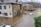 Severe flooding surrounds several residential buildings, submerging streets and partially covering cars. The water level is almost reaching the windows of the houses, and the current is visible, indicating strong water flow. Trees without leaves are visible in the background, suggesting it's a cold season.
