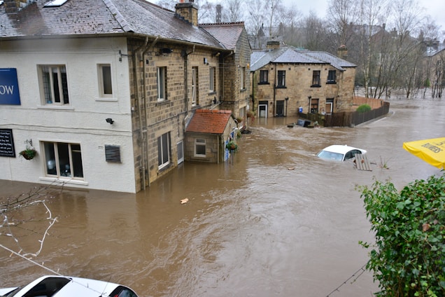 Severe flooding surrounds several residential buildings, submerging streets and partially covering cars. The water level is almost reaching the windows of the houses, and the current is visible, indicating strong water flow. Trees without leaves are visible in the background, suggesting it's a cold season.