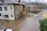 Severe flooding surrounds several residential buildings, submerging streets and partially covering cars. The water level is almost reaching the windows of the houses, and the current is visible, indicating strong water flow. Trees without leaves are visible in the background, suggesting it's a cold season.