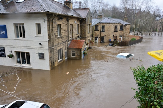 Severe flooding surrounds several residential buildings, submerging streets and partially covering cars. The water level is almost reaching the windows of the houses, and the current is visible, indicating strong water flow. Trees without leaves are visible in the background, suggesting it's a cold season.