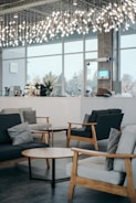 A modern indoor space featuring stylish wooden furniture, including sofas with cushions and round wooden coffee tables. The room is illuminated by overhead decorative lighting with hanging lights. Large windows allow natural light to enter, contributing to a bright and airy ambiance. In the background, there is a counter with various items and decorative plants.