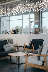 A modern indoor space featuring stylish wooden furniture, including sofas with cushions and round wooden coffee tables. The room is illuminated by overhead decorative lighting with hanging lights. Large windows allow natural light to enter, contributing to a bright and airy ambiance. In the background, there is a counter with various items and decorative plants.