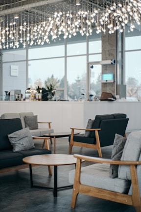A modern indoor space featuring stylish wooden furniture, including sofas with cushions and round wooden coffee tables. The room is illuminated by overhead decorative lighting with hanging lights. Large windows allow natural light to enter, contributing to a bright and airy ambiance. In the background, there is a counter with various items and decorative plants.