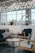 A modern indoor space featuring stylish wooden furniture, including sofas with cushions and round wooden coffee tables. The room is illuminated by overhead decorative lighting with hanging lights. Large windows allow natural light to enter, contributing to a bright and airy ambiance. In the background, there is a counter with various items and decorative plants.