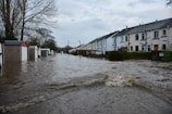Floodwaters cover a residential street, partially submerging garages and houses on either side. Trees without leaves are visible, and the sky is overcast, adding to the somber atmosphere.
