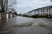 Floodwaters cover a residential street, partially submerging garages and houses on either side. Trees without leaves are visible, and the sky is overcast, adding to the somber atmosphere.
